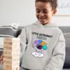 Kid wearing a grey melange hoodie featuring the 'Little Astronaut' design with custom name and playful space elements. The child is happily playing outdoors while showcasing the personalized astronaut-themed hoodie.