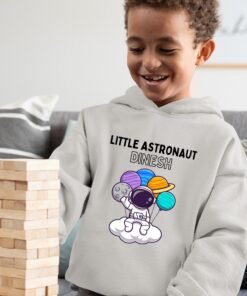 Kid wearing a grey melange hoodie featuring the 'Little Astronaut' design with custom name and playful space elements. The child is happily playing outdoors while showcasing the personalized astronaut-themed hoodie.